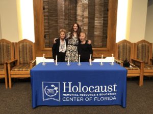 Three women standing in front of 7 lit candles that are on a table with a tablecloth that says Holocaust Memorial Resource & Education Center of Florida
